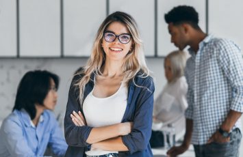 Confident blonde female manager posing with smile after conference with other employees. Asian programmer talking with african freelancer while fair-haired secretary laughing on foreground.. Confident blonde female manager posing with smile after conference with other employees. Asian programmer talking with african freelancer while fair-haired secretary laughing on foreground.